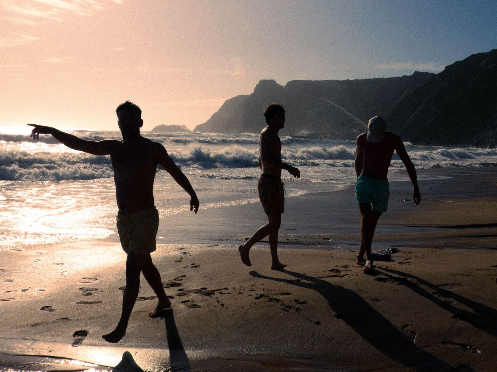 Three friends walking along a beach at sunset beneath coastal mountains